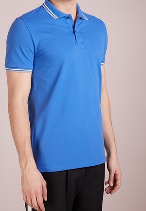 Sininen poolopaita, joka on valmistettu sileästä kankaasta. Paitassa on klassinen kaulus, jossa on valkoiset raidat, lyhyet hihat, mittatilaustyönä valmistettu istuvuus ja hienovaraisia ompeluyksityiskohtia.