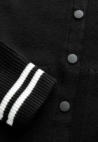 Zwarte cardigan met een ribbelmanchet met zwart-witte strepen en zwarte knopen aan de voorkant. Zachte textuur met een breiwerkontwerp.