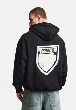 Mężczyzna w czarnej bluzie z kapturem z białym logo w kształcie tarczy i napisem „PROSTO” na plecach, w połączeniu z jasnoniebieskimi jeansami.