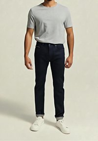 Homme portant un t-shirt gris clair à manches courtes, un jean foncé retroussé et des baskets blanches basses, debout sur un fond uni.