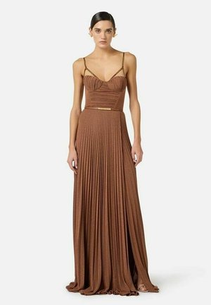 Donna che indossa un abito lungo marrone a pieghe con spalline sottili e un corpetto aderente arricciato, in piedi su uno sfondo bianco.