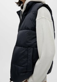 Svart puffväst med hög krage och dragkedja, prydd med horisontell sömnad och en sidoficka. Bärs över en ljusgrå hoodie.