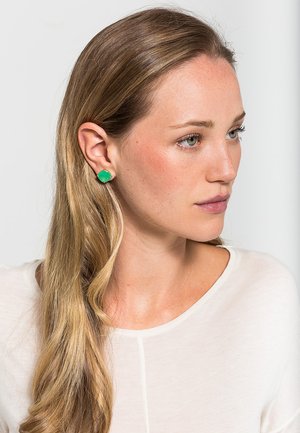 Grüne Steinearrings in runder Form, vor einem neutralen Hintergrund. Das Modell trägt ein weißes Oberteil, das die Details der Ohrringe hervorhebt.