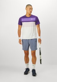 Lilla og hvit sportsskjorte med horisontale fargeblokker, parret med lyseblå shorts. Iført treningesko og holder en tennisracket.
