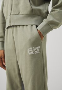 Sportski trenirku, maslinasto zelena tkanina, rebrasti rukavi i pojas, s izvezenim EA7 logom u bijeloj boji na lijevoj nozi.