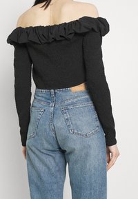 Top nero off-the-shoulder con scollo arricciato e tessuto texturizzato, abbinato a jeans blu a vita alta con etichetta in pelle sul retro.