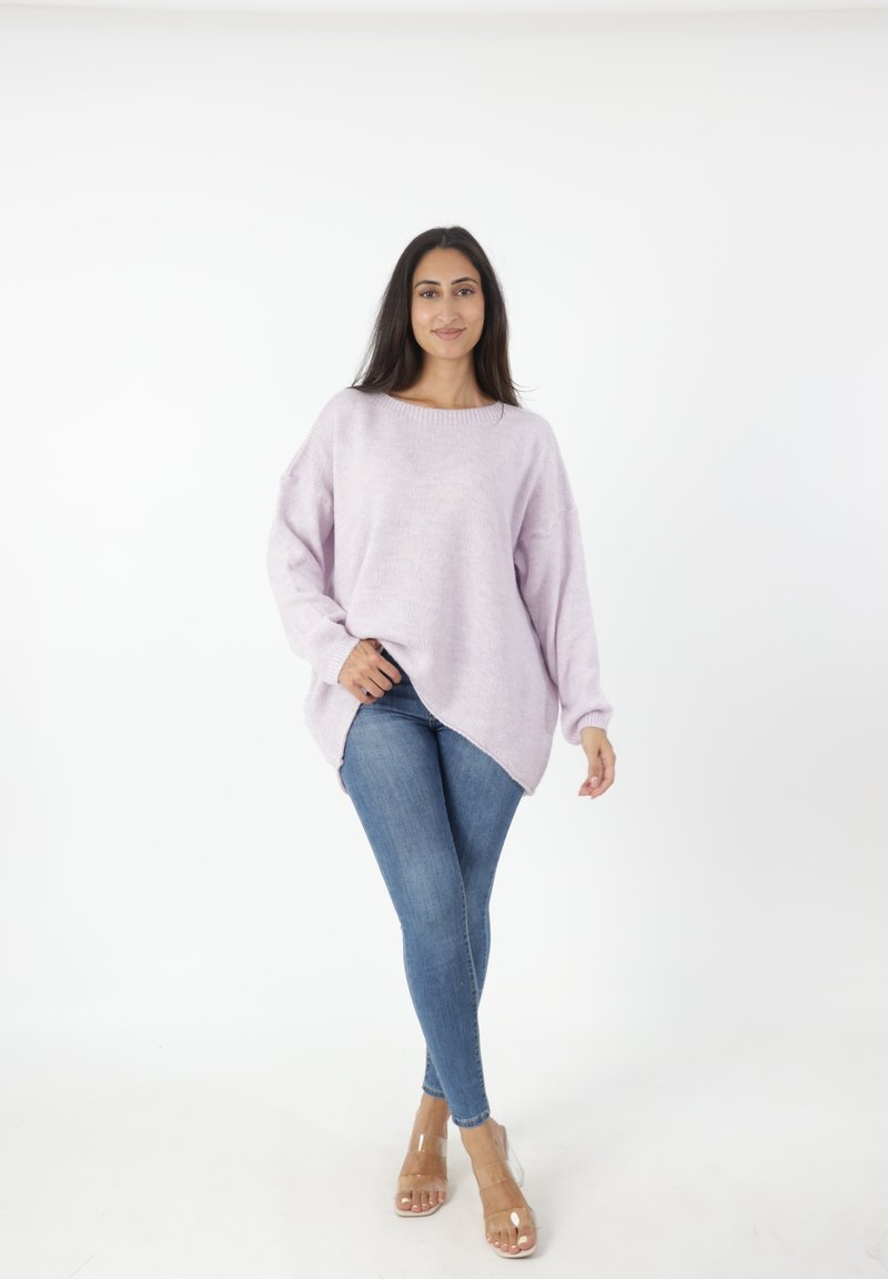 Suéter oversized de color púrpura claro con escote barco y aberturas laterales, combinado con jeans ajustados azules y sandalias de tacón transparentes.