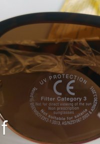 Gafas de sol marrones con un marco oscuro. La lente muestra detalles de protección UV y marcas de categoría de filtro. Película protectora transparente visible.