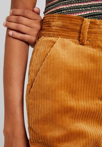 Kordbyxor i en varm, bärnstensfärg, med bred, vertikal ribbning och midjeband. Sidfickor och jämn textur synliga.