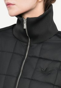 Casaco puffer preto com colarinho alto, apresentando um fecho frontal e padrão acolchoado. Logótipo bordado no lado esquerdo do peito.