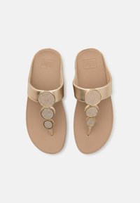 Gullige sandaler med en bred stropp som har tre sirkulære dekorasjoner. Tan fotseng med teksturelle detaljer. Flatt såledesign.