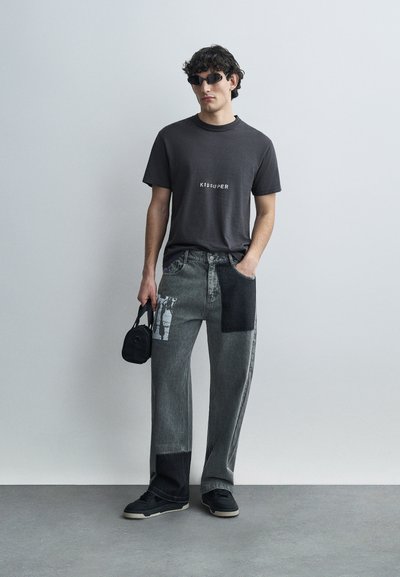 Μαύρο γραφικό t-shirt με λευκό κείμενο; τζιν γκρι πατς με ποικίλες υφές; μαύρα αξεσουάρ; μαύρα αθλητικά παπούτσια.