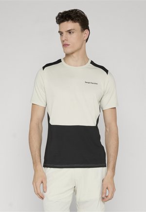 Mand iført kortærmet, todelt beige og sort Sergio Tacchini t-shirt stående mod en ensfarvet baggrund.