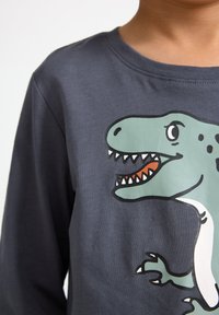 Child wearing a dark gray long-sleeve shirt with a smiling green cartoon dinosaur graphic on the front.
