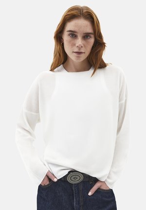 Junge Frau mit roten Haaren und Sommersprossen, die einen weißen Pullover und dunkle Jeans mit einer dekorativen Gürtelschnalle trägt, Hände in den Taschen.