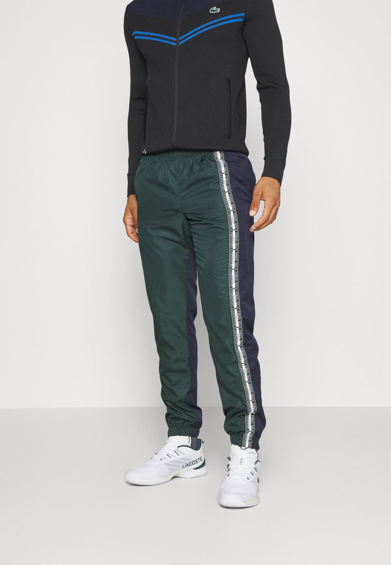 Pantalons de jogging verts et bleu marine avec une taille élastique, featuring une bande logo blanche sur le côté. Assortis avec des chaussures de sport blanches.