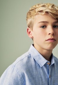 Lyseblå skjorte med knapper lavet af tekstureret stof, med krave og en træknap, båret af en ung person med kort blondt hår.