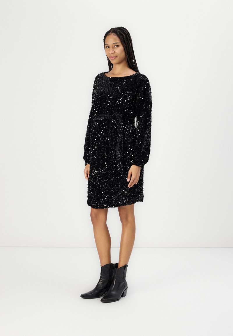 Vestido preto com lantejoulas, de corte solto, mangas longas, decote curvo e detalhe de cintura atada. Usado com botas pretas de tornozelo.