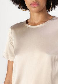 Beige selyem rövid ujjú felső, kerek nyakkivágással, sima textúrával és finom fényű felülettel. Egyszerű dizájn, minta vagy díszítés nélkül.