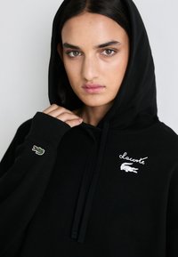Juodas džemperis su raišteliu, su išsiuvinėtu baltu Lacoste logotipu ir mažo žalio krokodilo akcentu ant rankovės. Minkšta medžiagos tekstūra.