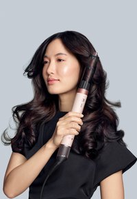 Strumento per acconciature con manico rosa, cilindro arricciacapelli nero, impugnatura testurizzata e pulsanti. Una donna lo tiene mentre mostra i suoi capelli voluminosi.