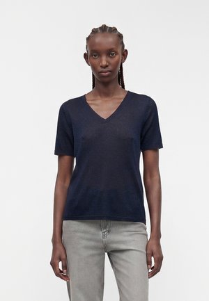 Femme aux cheveux tressés portant un haut bleu marine à manches courtes et col en V ainsi qu'un jean gris clair, debout devant un fond blanc uni.