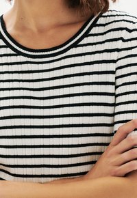 Top blanco a rayas con finas rayas negras, textura acanalada y cuello redondo. Hombros visibles, manos cruzadas en la cintura.