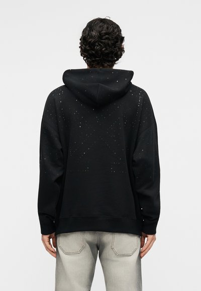 Cilvēks ar cirtainiem matiem, tērpies melnā kapučjakā ar smalku punktveida zvaigžņu rakstu uz muguras un gaiši pelēkās džinsu biksēs, stāv ar muguru pret skatītāju.