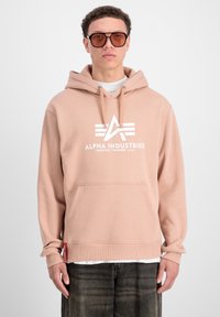 Šviesiai rožinė pūkinė striukė su kengūros kišene ir stringų kapu; priekyje yra balta "ALPHA INDUSTRIES" logo. Pagaminta iš medvilnės.
