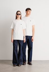 Λευκά κοντομάνικα μπλουζάκια σε συνδυασμό με σκούρα μπλε τζιν φαρδιάς γραμμής. Τα μπλουζάκια έχουν ένα μικρό λογότυπο στην αριστερή πλευρά του στήθους. Ομαλή υφή υφάσματος.