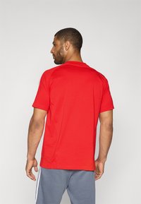 Red athletic t-shirt with short sleeves, round neckline, smooth texture, and contrasting white side stripes, worn with gray shorts.