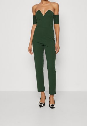 Green, off-the-shoulder jumpsuit with a deep V-neck, slim fit, and ankle-length legs. Worn with black heels featuring decorative buckles.