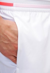 Main reposant dans la poche d’un short de sport blanc avec une ceinture élastique rose et rouge.