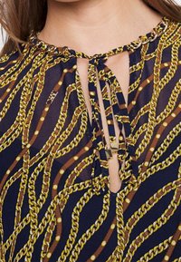 Laivastonsininen pusero, jossa on ketju- ja helmiraita, mukana nyörityksellä ja avainaukko-kaula-aukolla. Valmistettu kevyestä kankaasta, jolla on sileä rakenne.