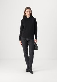 Sweat-shirt à capuche noir avec un logo discret, associé à un jean gris foncé et des bottines noires; le modèle tient un petit sac à main noir.