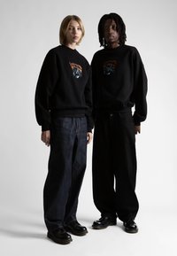 Felpe nere con stampa grafica, abbinate a jeans scuri a vestibilità ampia e pantaloni neri. I modelli indossano scarpe nere con suole spesse.