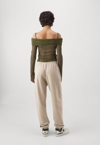 Olivgrünes Off-Shoulder-Oberteil mit strukturierten Details, kombiniert mit hellbeigen Jogginghosen. Model trägt weiße und grüne Sneakers.