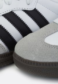 Zapatilla de cuero blanca con tres rayas negras, puntera de gamuza gris y suela de goma negra texturizada. Presenta detalles cosidos y cordones.