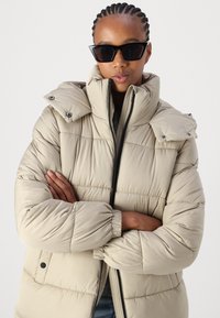 Beige puffjakke med høy krage, trykknapper på skuldrene, quiltet design og elastiske mansjetter. Brukt med svarte solbriller.