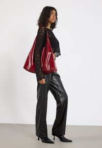 Femme portant un haut en dentelle noire et un pantalon en cuir, portant un grand sac à main rouge brillant sur son épaule, se tenant de côté sur un sol gris.