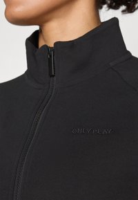 Chaqueta negra con cremallera que presenta un cuello alto y un tejido texturizado, con un logo en relieve "ONLY PLAY" en la parte delantera cerca de la cremallera.
