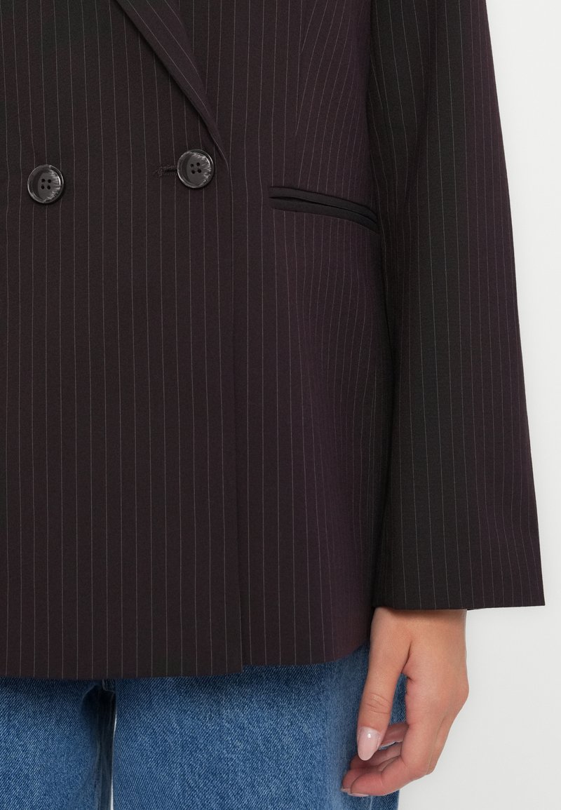 Blazer doppiopetto in tessuto scuro con sottili righe verticali, caratterizzato da due bottoni neri e una tasca sul petto. Maniche lunghe con taglio dritto.