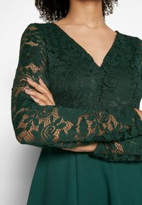 Dunkelgrünes Kleid mit V-Ausschnitt und langen Ärmeln aus floraler Spitze. Der Stoff hat eine glatte Oberfläche mit Kanten, die das Spitzenmuster zur Geltung bringen.