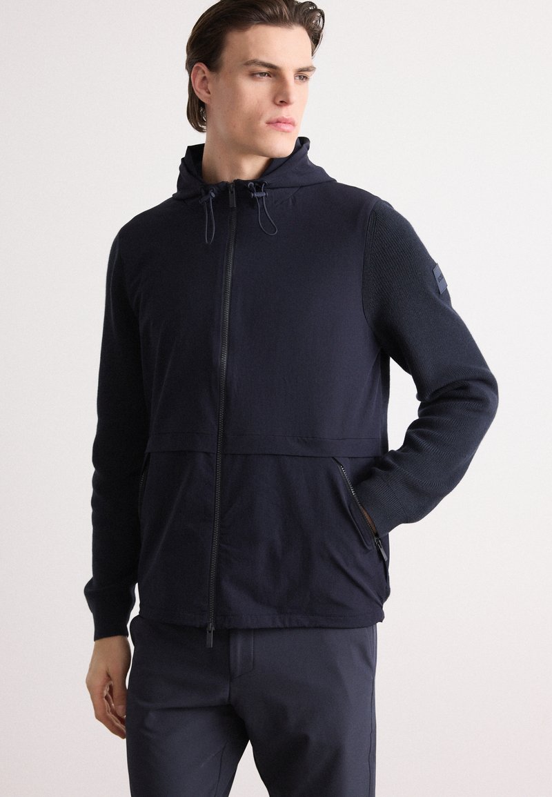 Navy zip-up jacket with a hood, textured knit sleeves, two side pockets, and adjustable drawstrings for a secure fit.
