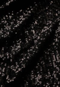 Tissu noir recouvert de petites paillettes brillantes. La surface est texturée et capte la lumière, créant un effet scintillant.