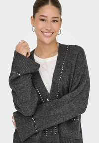 Šedý pletený kardigan s hlubokým výstřihem do V, dlouhými rukávy, zapínáním na knoflíky a dekorativními nýty podél okrajů. Měkká struktura a volný střih.