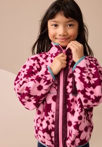 Rózsaszín polárdzseki lila virágmintával, teljes cipzárral, magas nyakkal és türkiz részletekkel. Puha textúra és kényelmes szabás.