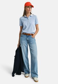 Helles blaues Poloshirt mit Kragen, kombiniert mit ausgebleichten Weitbein-Jeans. Eine orangefarbene Kappe, ein brauner Gürtel und weiße Sneaker runden den Look ab.