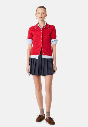 Femme portant un cardigan rouge à manches courtes par-dessus une chemise bleu clair, une jupe plissée à rayures bleu marine, des chaussures marron et des chaussettes beiges, debout sur fond blanc.