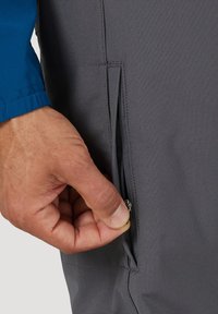 Hand mit blauem Ärmel, die den Reißverschluss an der grauen Hosentasche vor weißem Hintergrund öffnet.